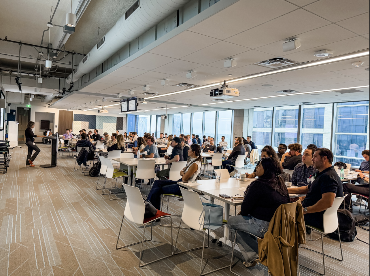 A large conference room filled with people attending a presentation, seated at white tables with some taking notes or listening, with floor-to-ceiling windows showing a cityscape outside.